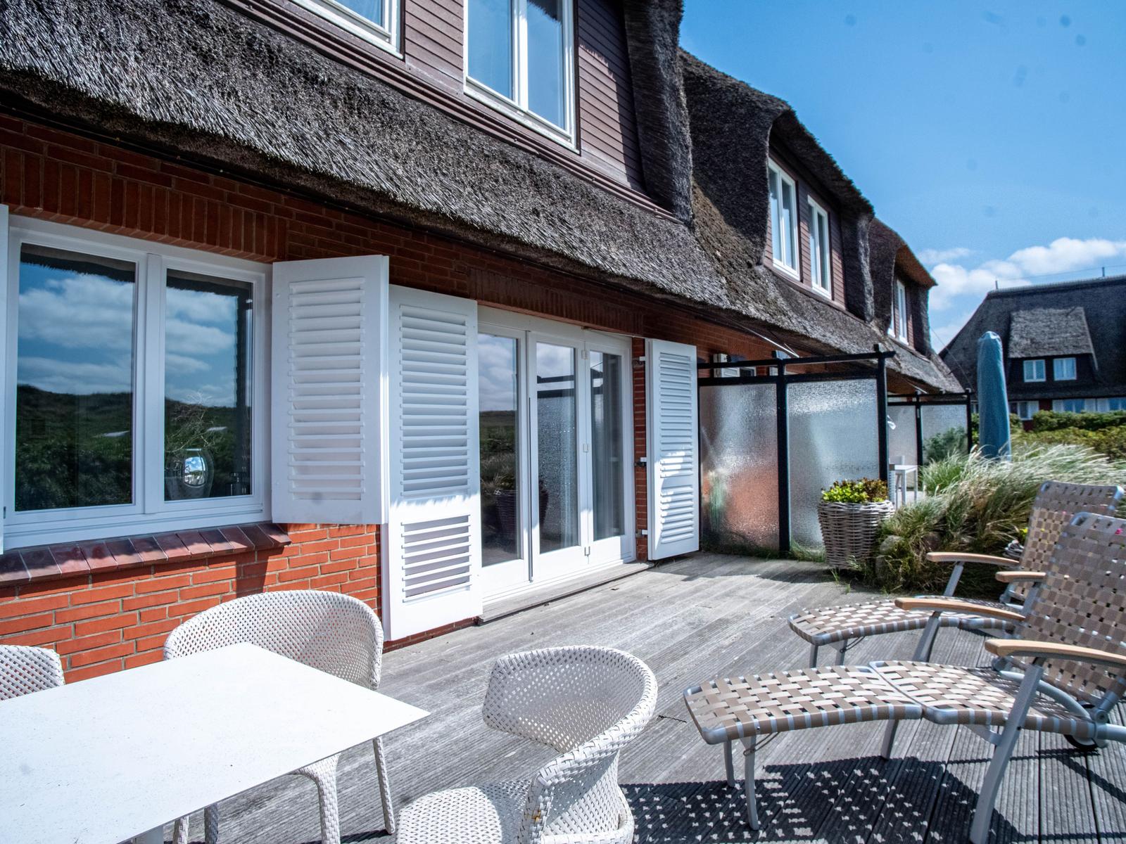 Terrasse mit Tisch, Stühlen und Liegestühlen vor einem Haus mit Schiebetüren und Dachreiter.