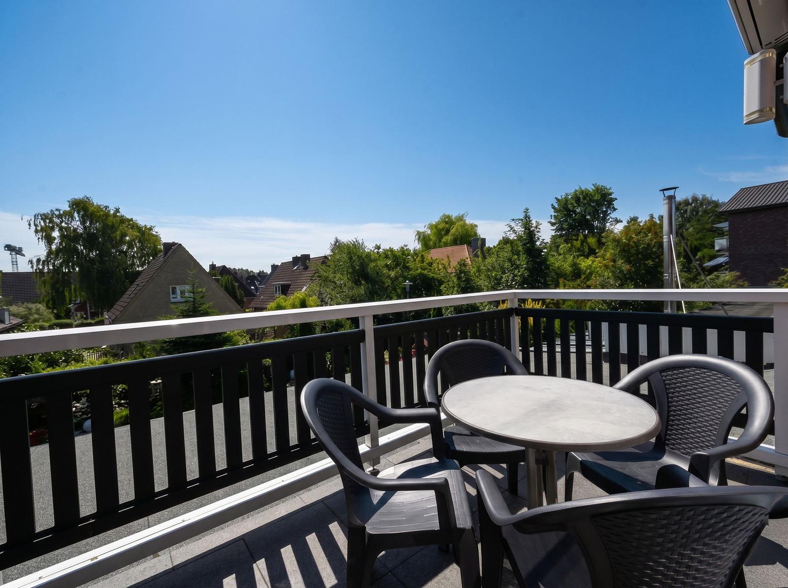 Terrasse mit Tisch und Stühlen, Blick auf Grün und Häuser unter blauem Himmel.