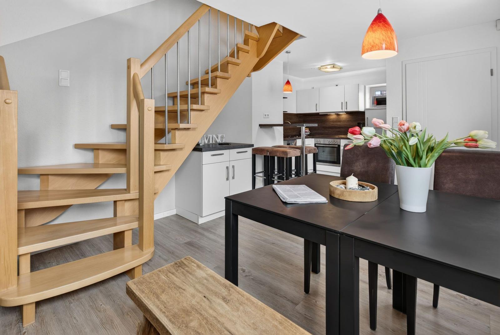 Essbereich mit Tisch, Stühlen und Blumen. Holztreppe führt zur Küche.