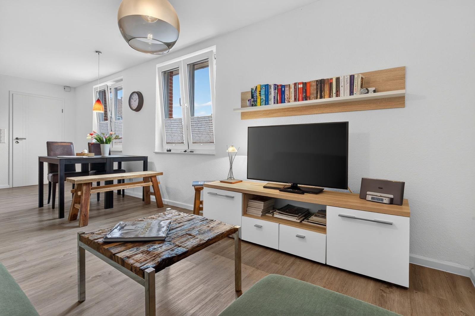 Wohnzimmer mit TV, Bücherregal und Esstisch. Holzboden und große Fenster.