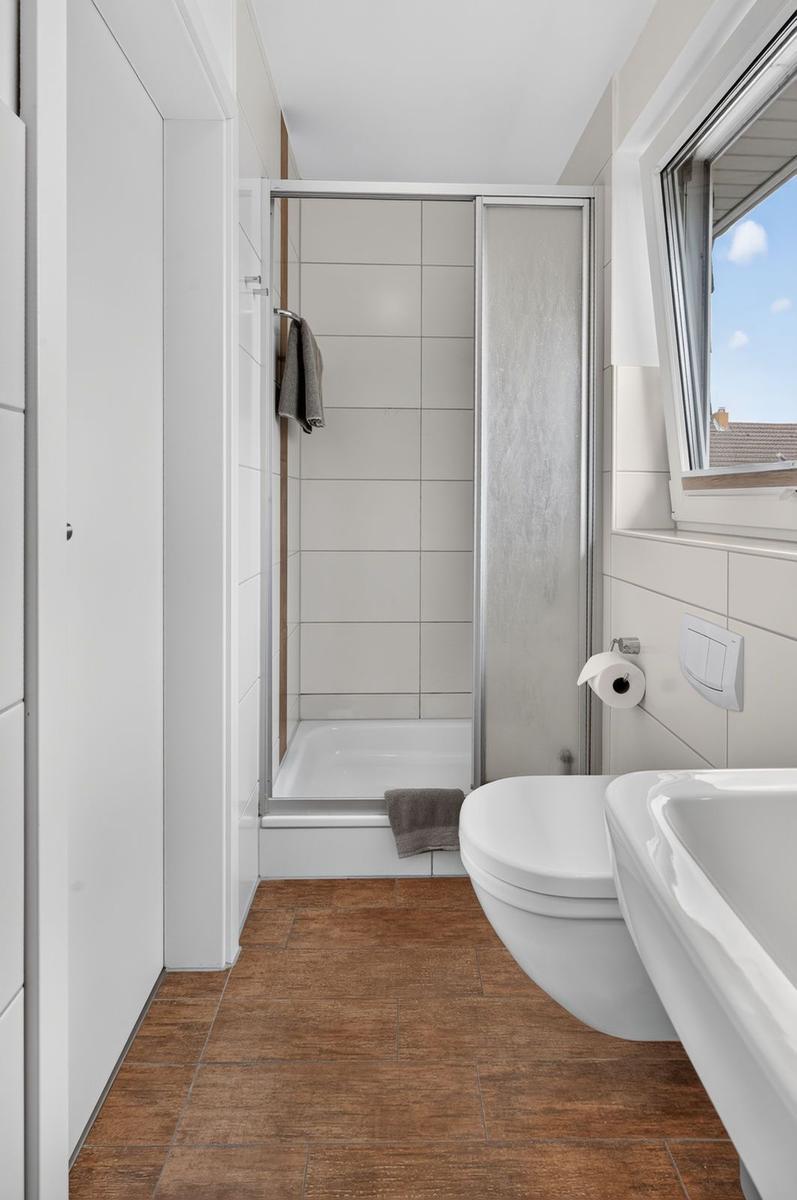 Badezimmer mit Dusche, Toilette und Waschbecken. Holzoptik Boden. Fenster mit Blick.