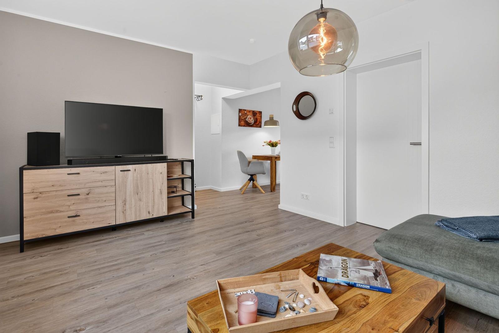 Wohnzimmer mit TV, Couch und Holztisch. Im Hintergrund ist ein Essbereich zu sehen.