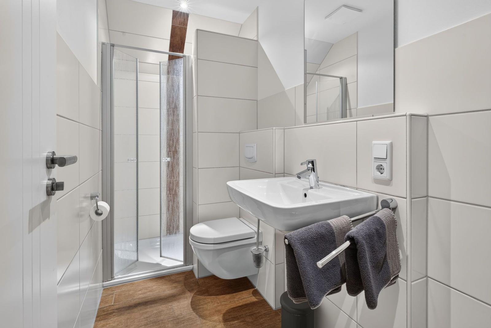 Badezimmer mit Dusche, Waschbecken und Toilette. Holzfußboden und weiße Fliesen.