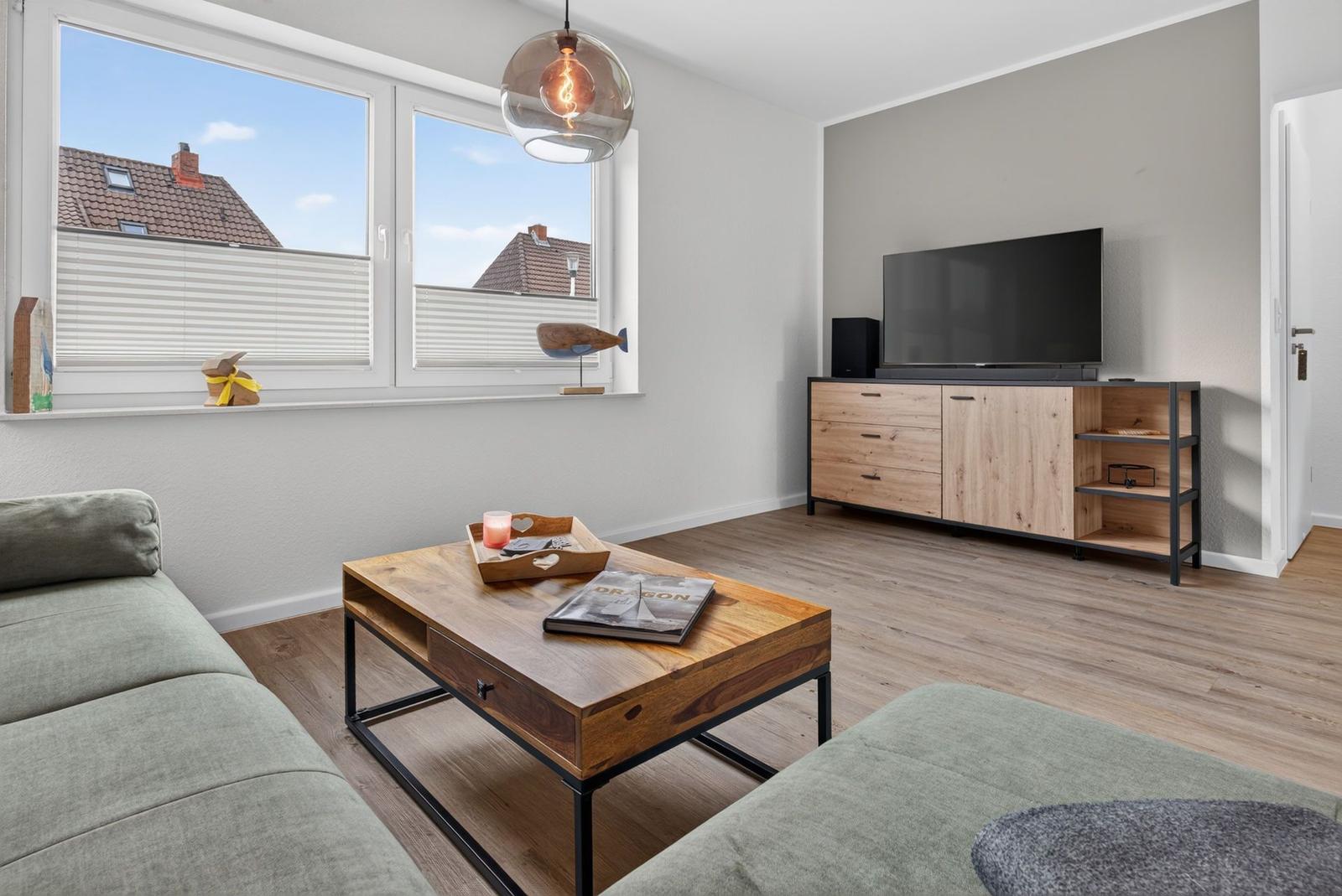Wohnzimmer mit Sofa, Couchtisch und Fernseher. Große Fenster mit Blick auf Dächer.