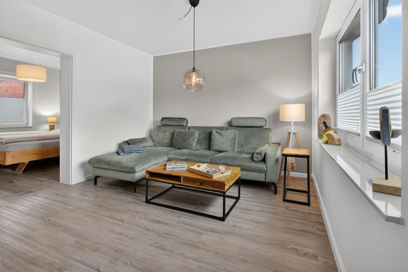Wohnzimmer mit grünem Sofa, Holzfußboden und großem Fenster. Blick in Schlafzimmer.