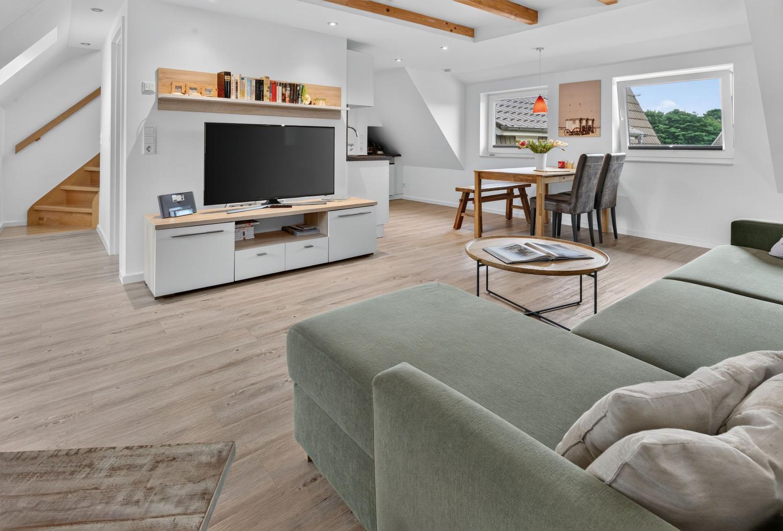 Wohn- und Essbereich mit Sofa, TV und Tisch. Holzboden und Fenster mit Blick.