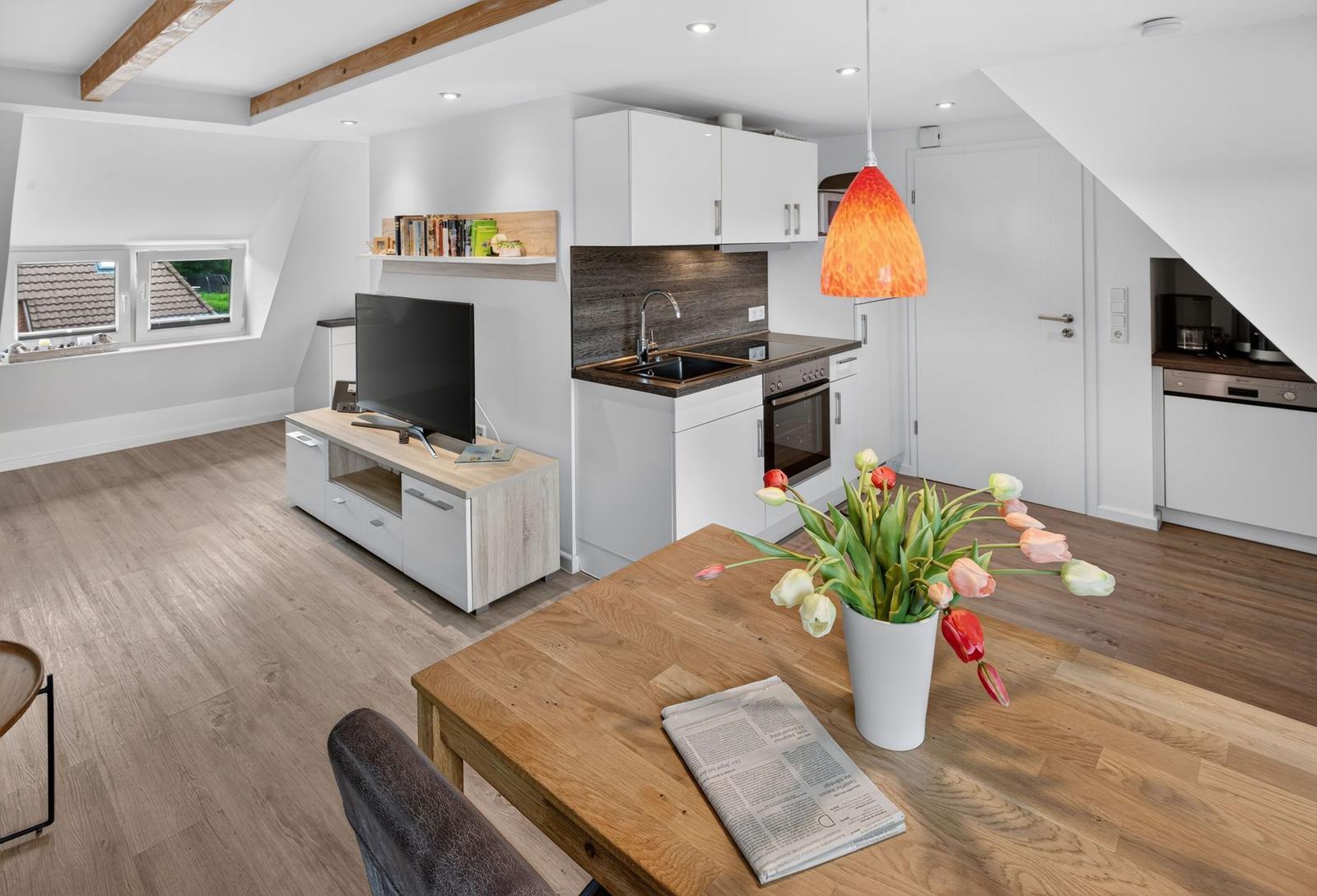 Wohnküche mit Esstisch, TV und Fenster. Holzboden und weiße Küchenzeile.