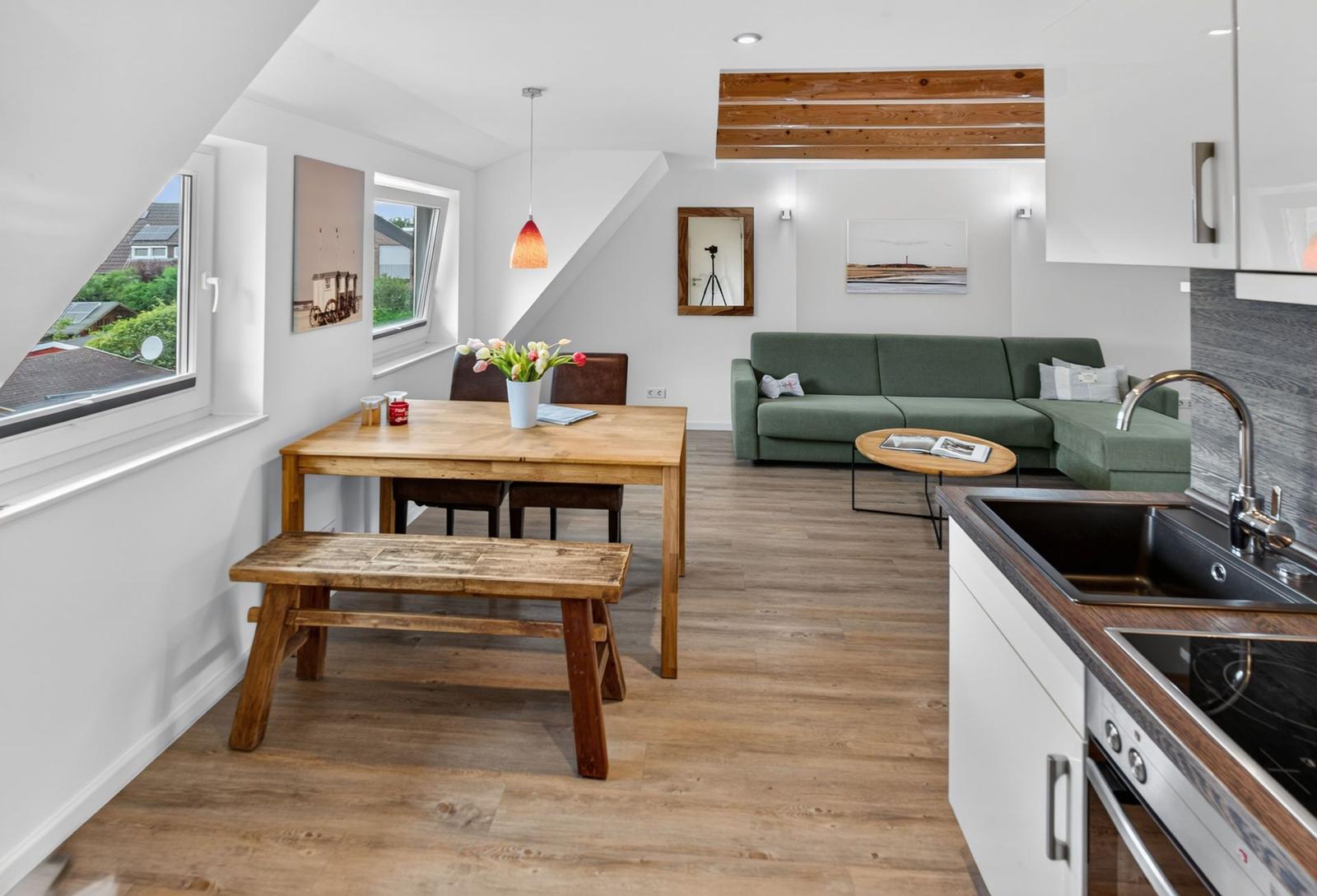 Wohnküche mit Essbereich und Sofa. Holzboden, Fenster und Küchenzeile sichtbar.