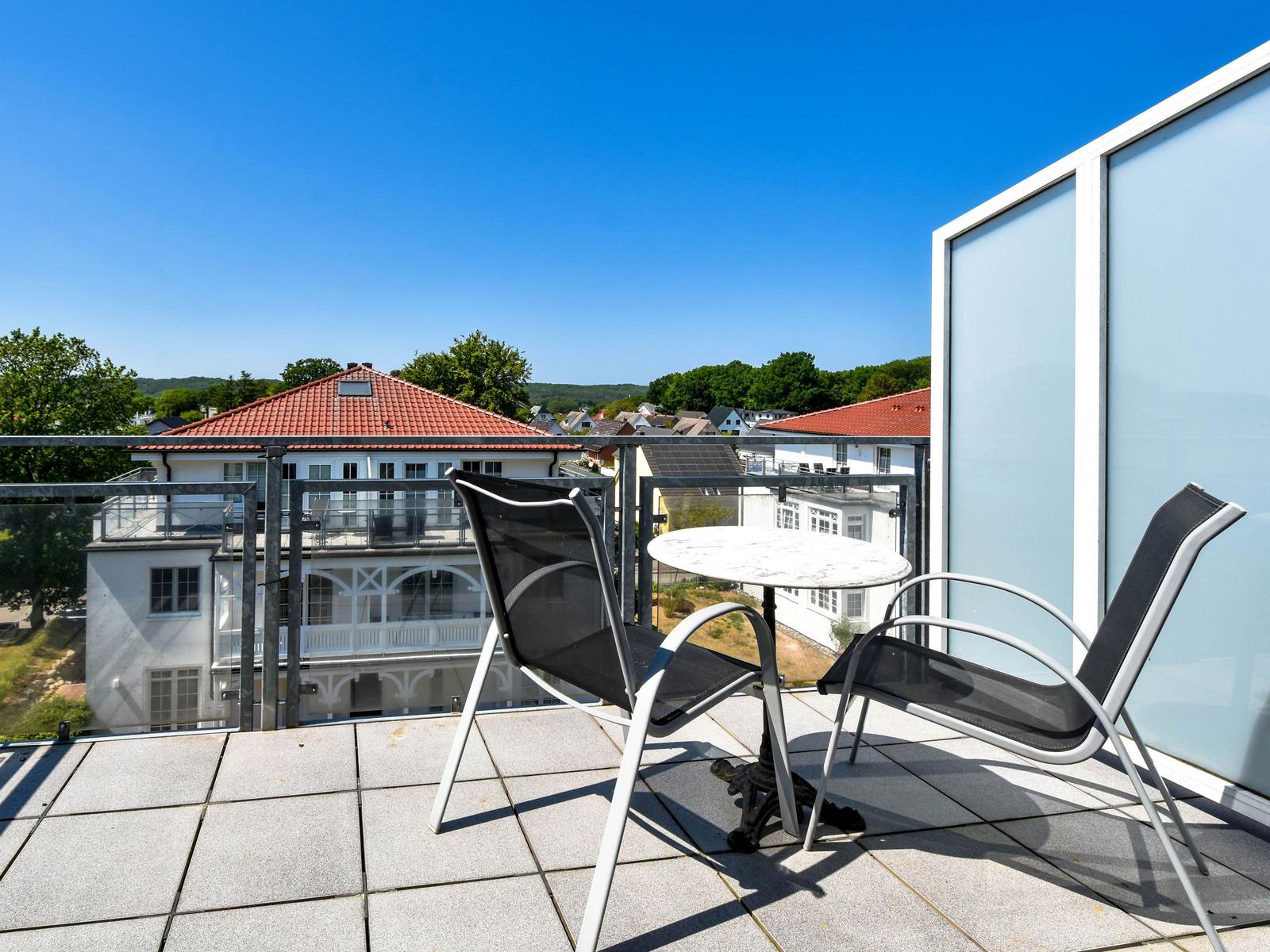 Terrasse mit Tisch und zwei Stühlen. Blick auf Wohnhäuser und grüne Hügel unter blauem Himmel.