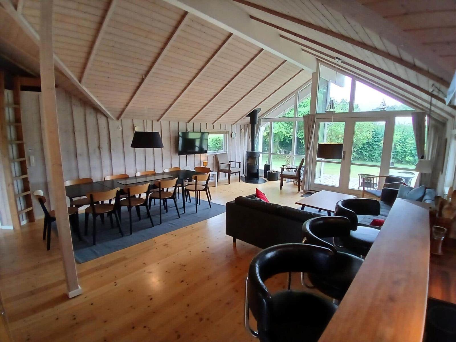 Ein Holzhaus mit Terrasse und Garten. Umgeben von Bäumen und Sträuchern unter blauem Himmel.
