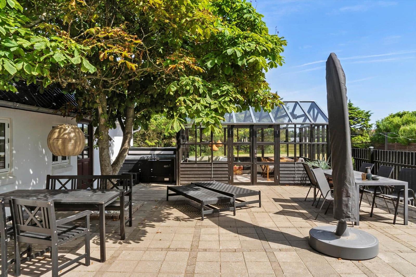 Glasveranda mit Sitzbereich und Tisch. Tür offen zur Terrasse. Grün hinter Glas.
