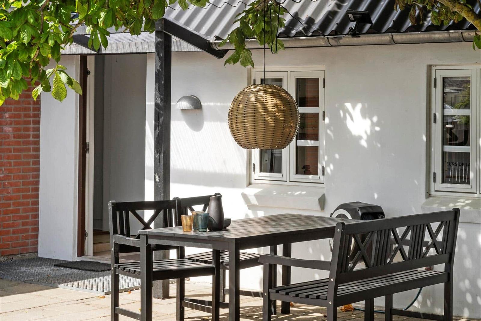 Terrasse mit Sitzgelegenheiten und Holztisch unter Glasdach. Hängelampe und Pflanzen sichtbar.