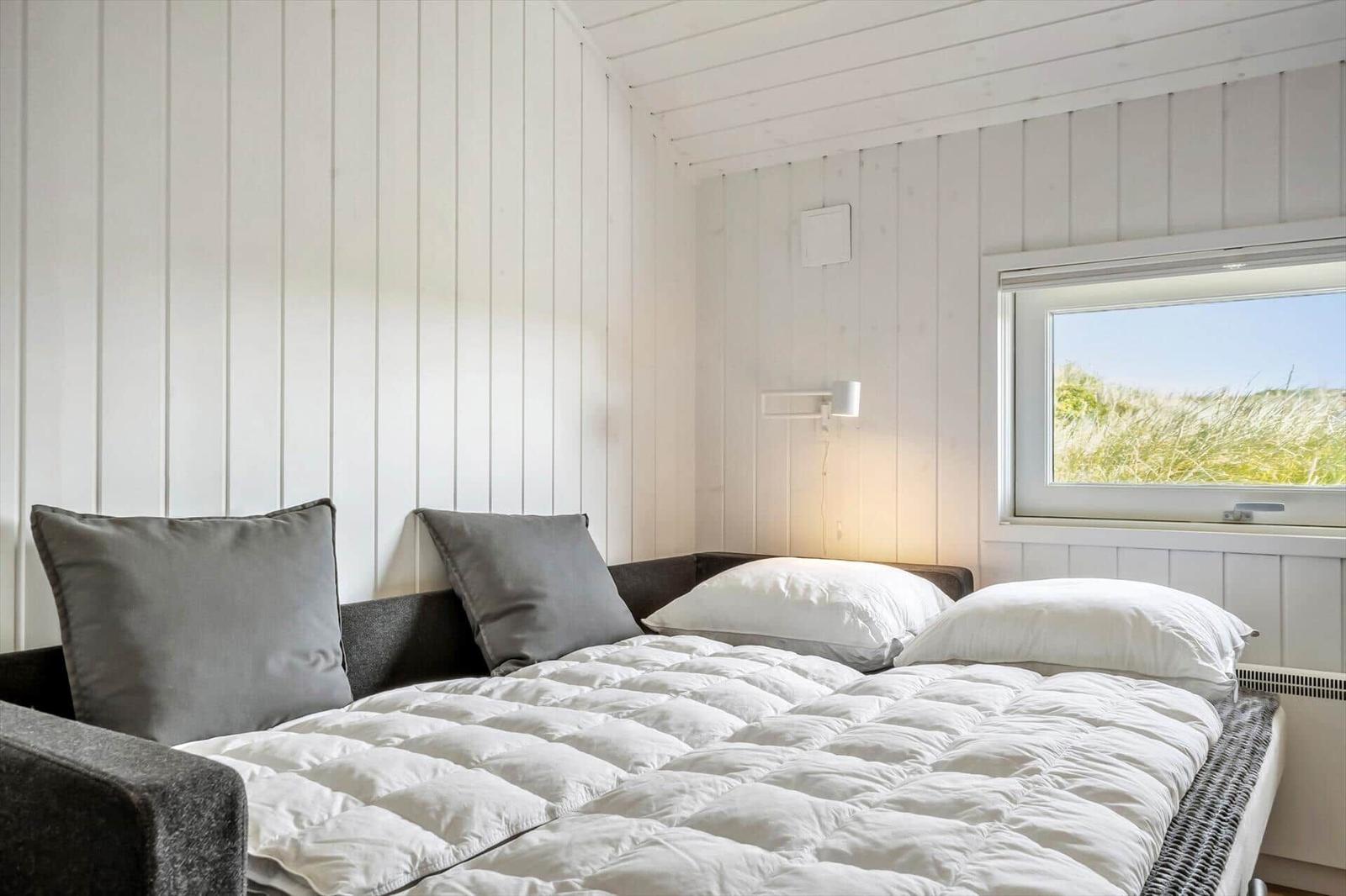 Schlafzimmer mit weißer Holzwand, Bett mit Kissen und Fensterblick auf Gras.