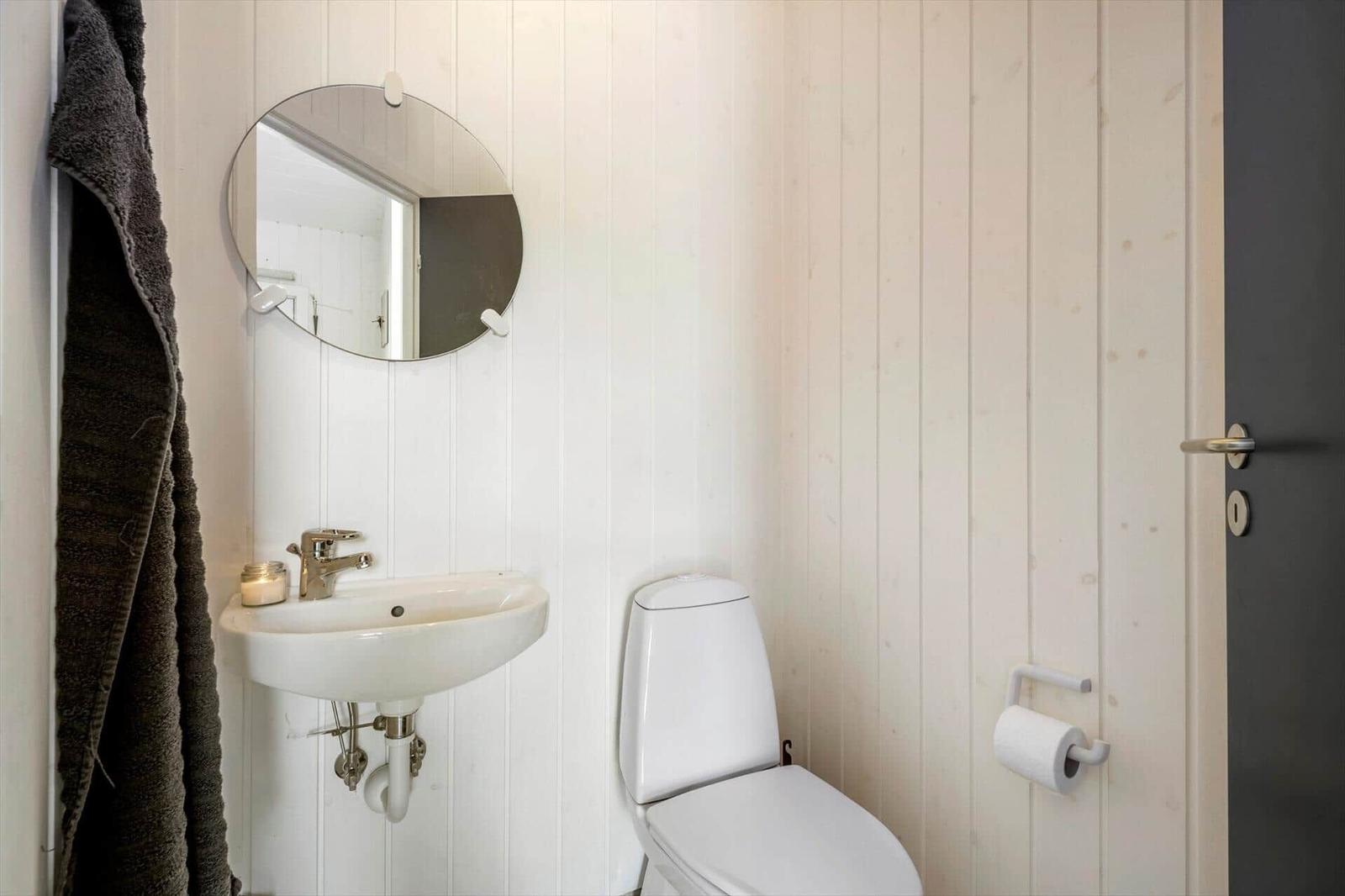 Badezimmer mit Toilette, Waschbecken und runden Spiegel an weißen Holzwänden.