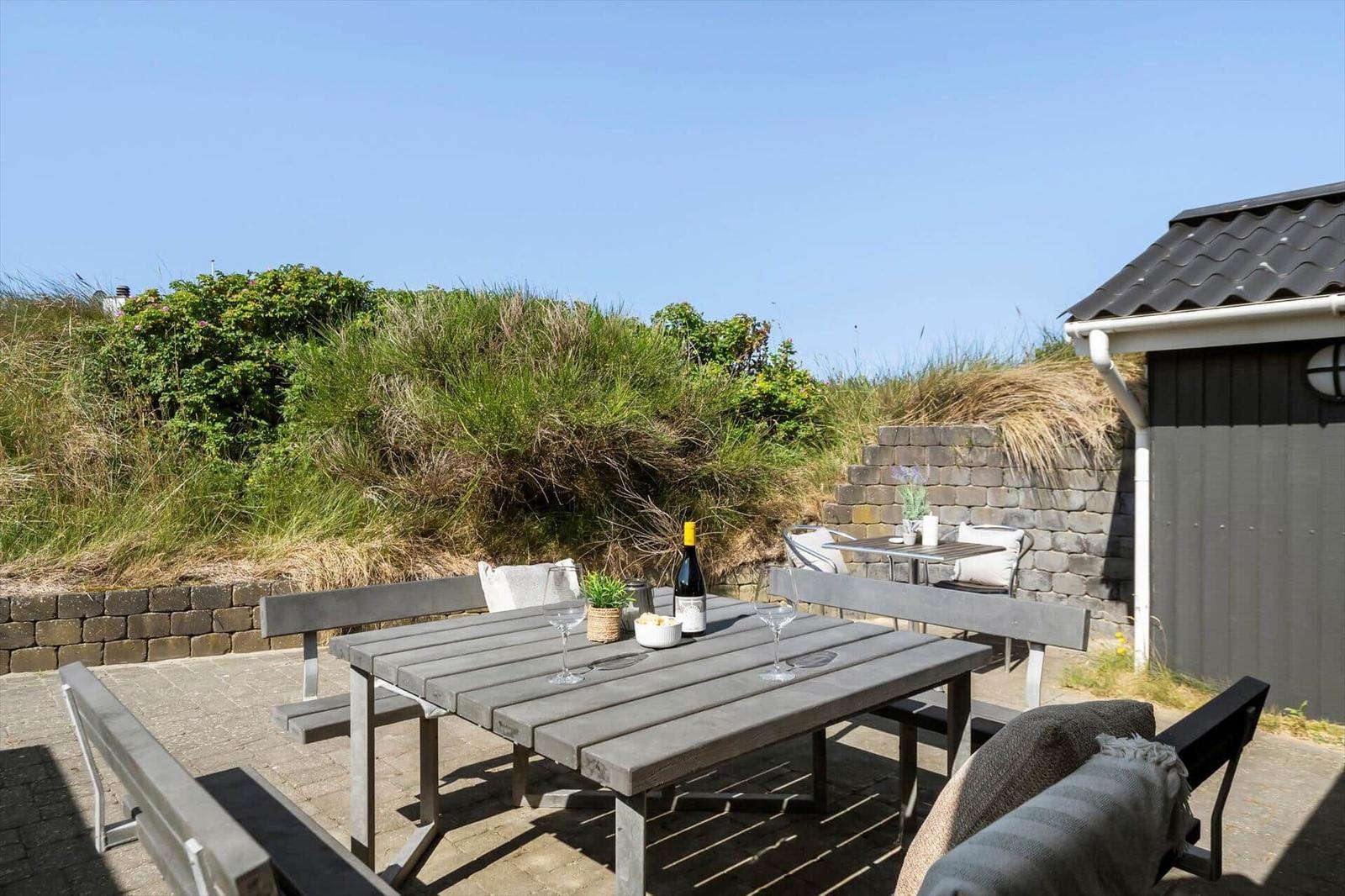 Terrasse mit Tisch, Bänken und Weinflasche vor Gras- und Steinmauer.