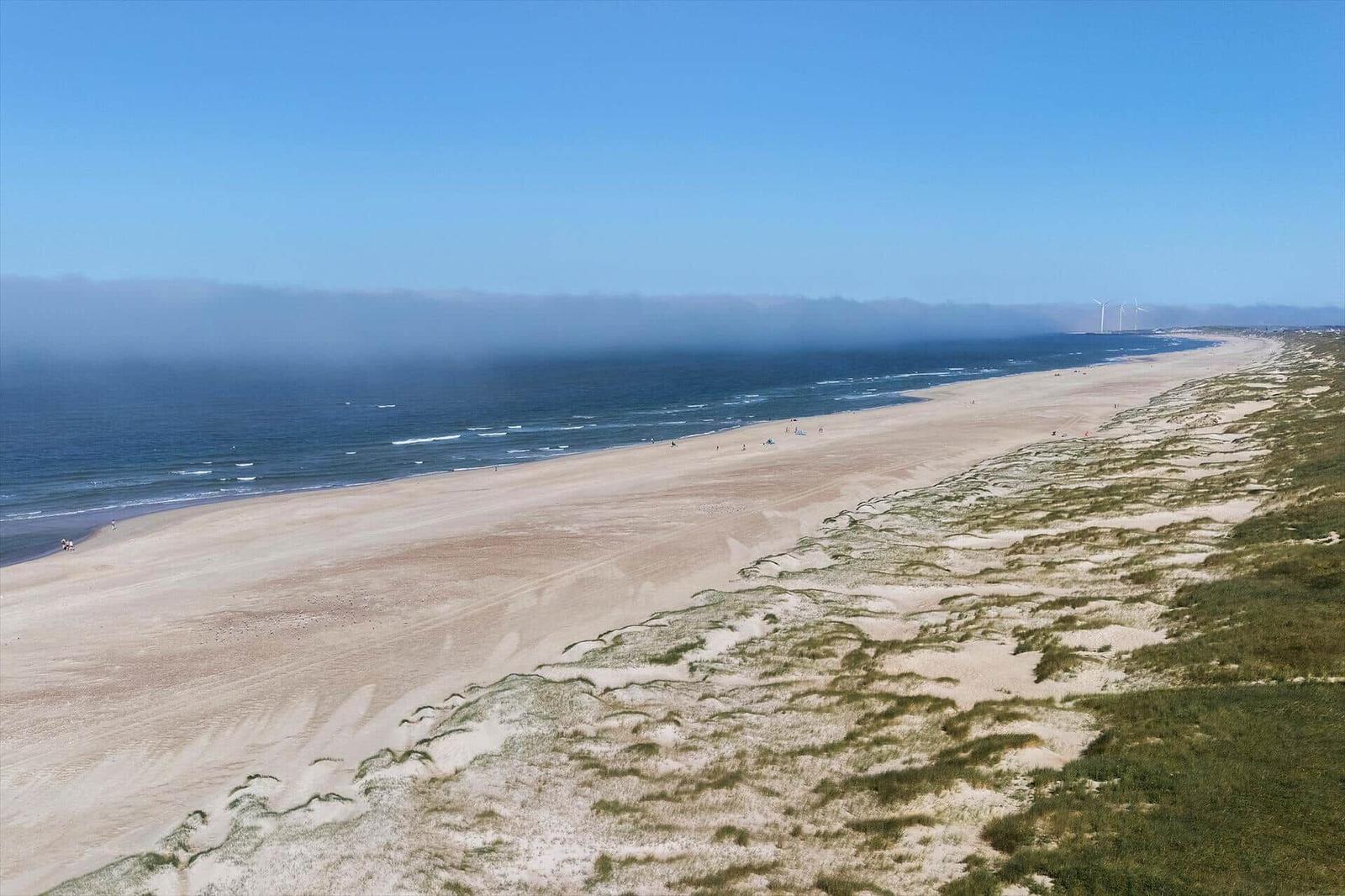 Weitläufiger Strand mit Dünen und Windkraftanlagen im Hintergrund.
