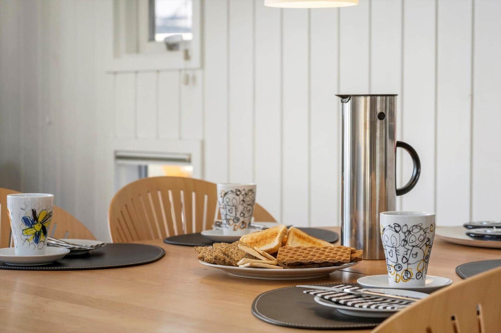 Esstisch mit Tassen, Kaffeebereiter und Gebäck auf dunklen Untersetzer.