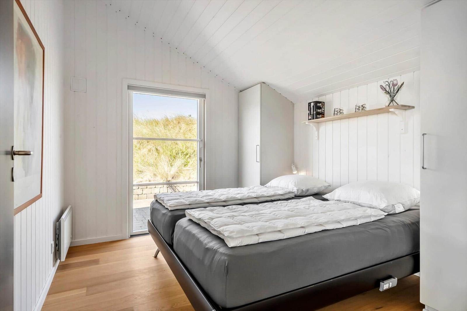 Schlafzimmer mit Doppelbett, weißer Wandverkleidung und Fenster zur Außenterrasse.