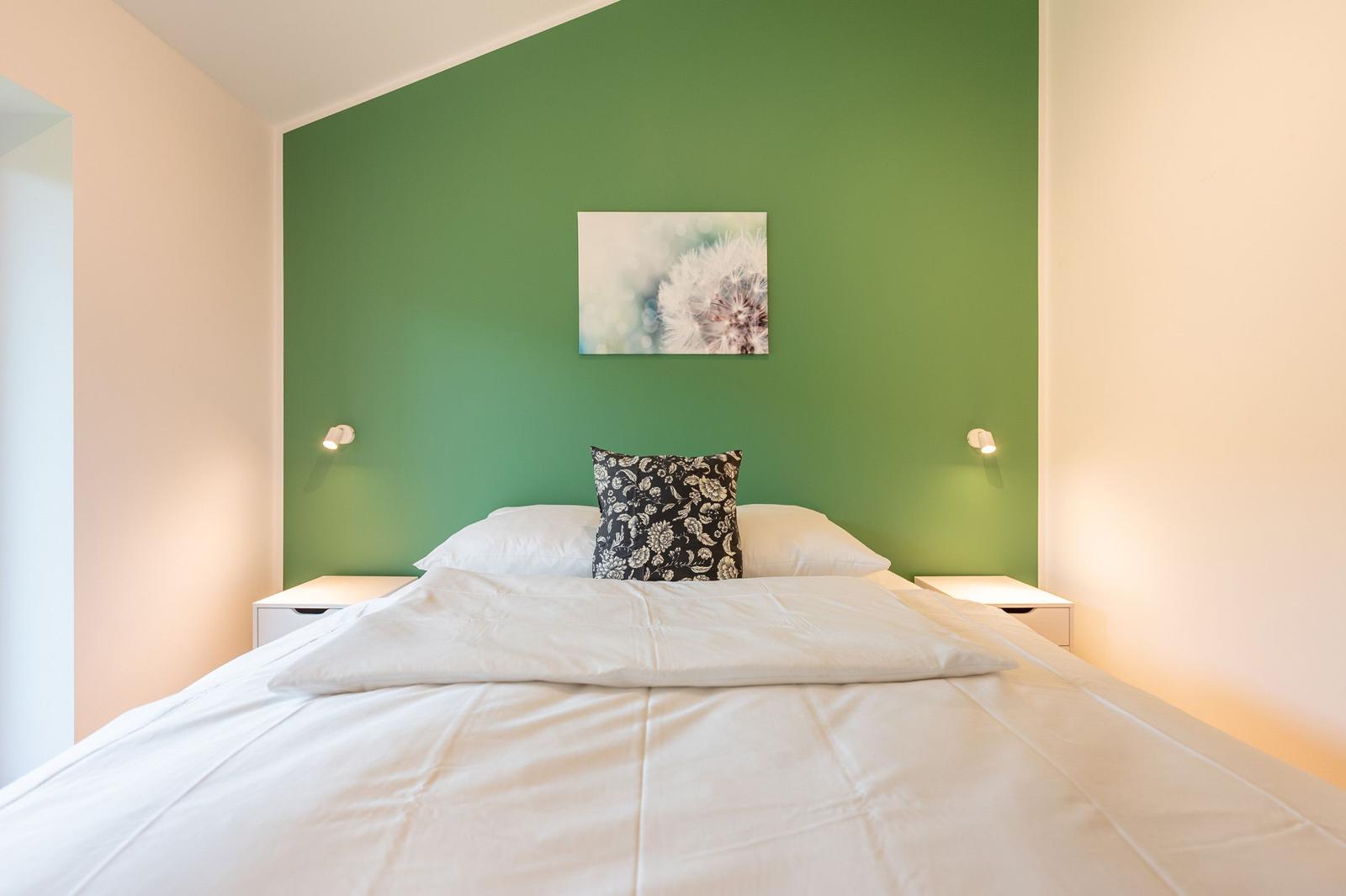 Schlafzimmer mit grüner Wand, Bett mit weißer Bettwäsche und Kissen, zwei Nachttische mit Lampen.