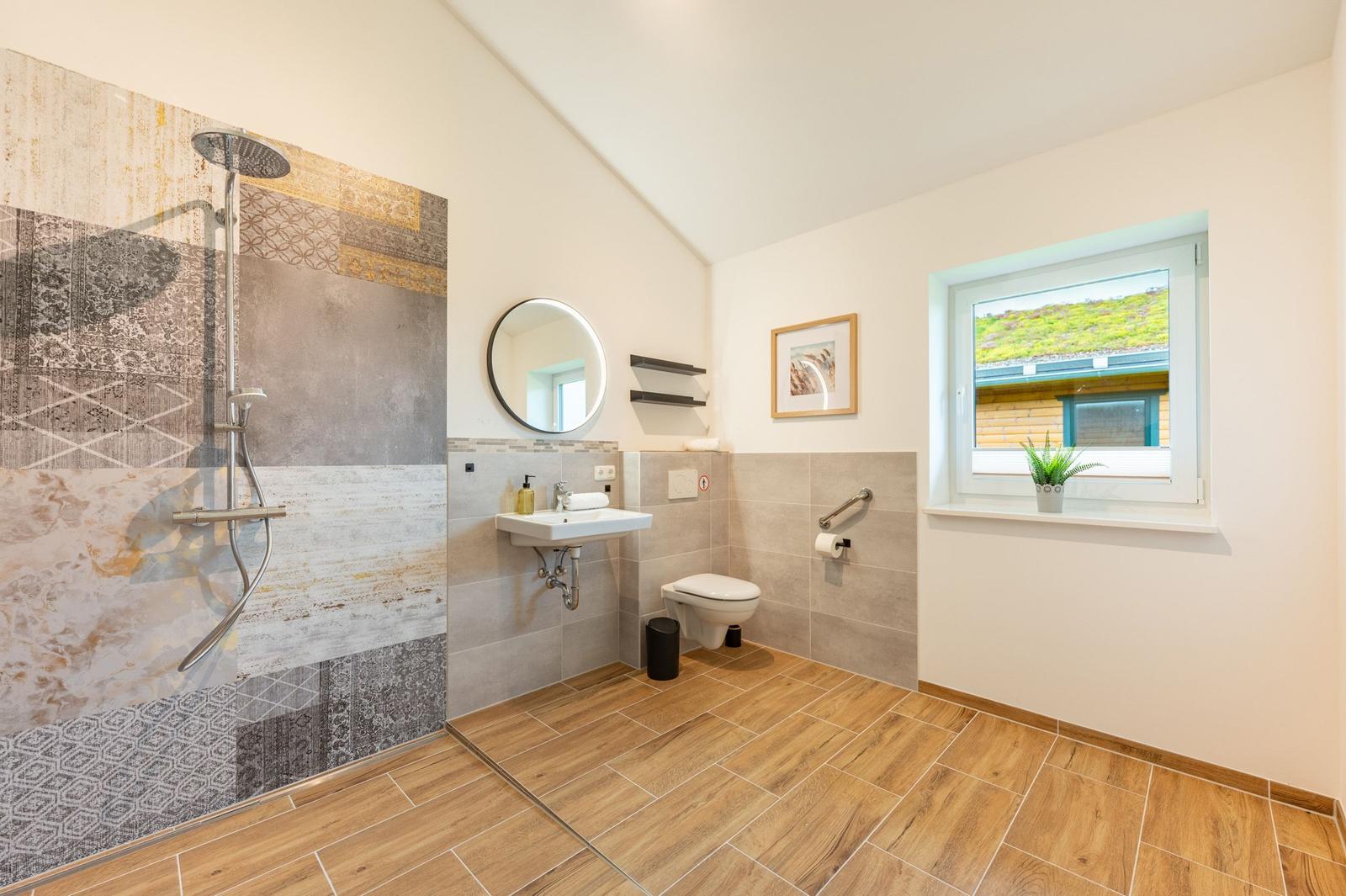 Badezimmer mit Dusche, Waschbecken und WC. Holzoptik Boden, Fenster mit Blick.
