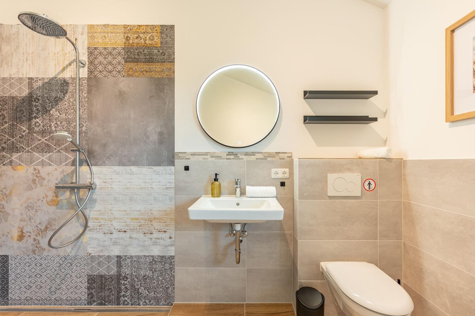 Badezimmer mit Dusche, Waschbecken und WC. Wand mit Mosaikfliesen und runder Spiegel.