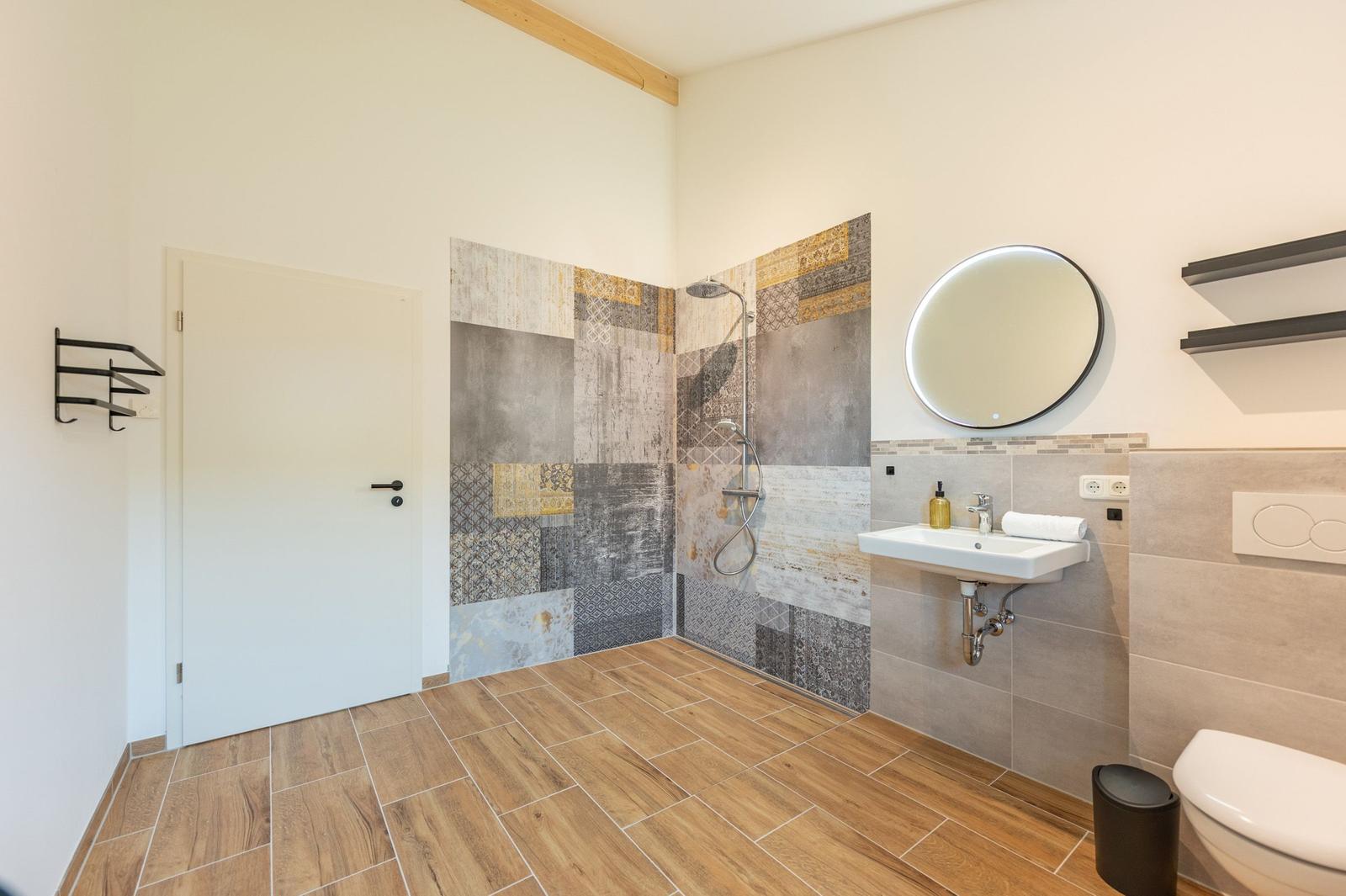 Bad mit Dusche, Waschbecken und Toilette. Fliesenwand und Holzdielenboden.
