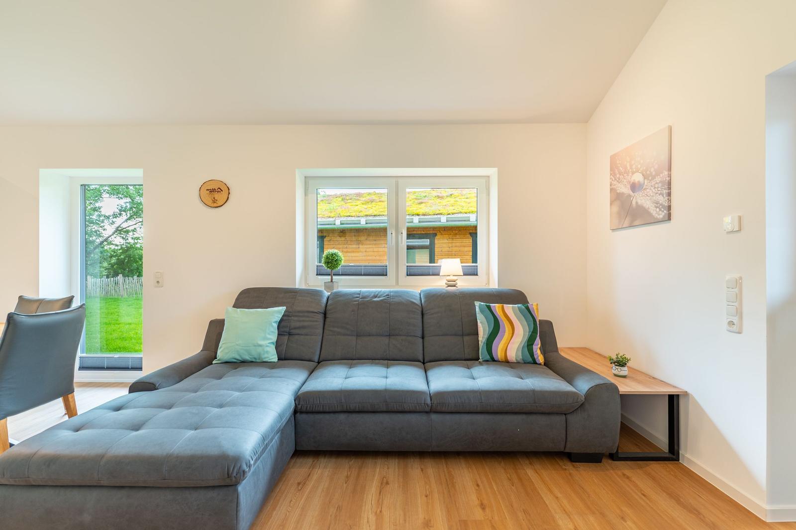 Ein Wohnzimmer mit grauem Sofa, Holzboden und Fenster zum Garten.