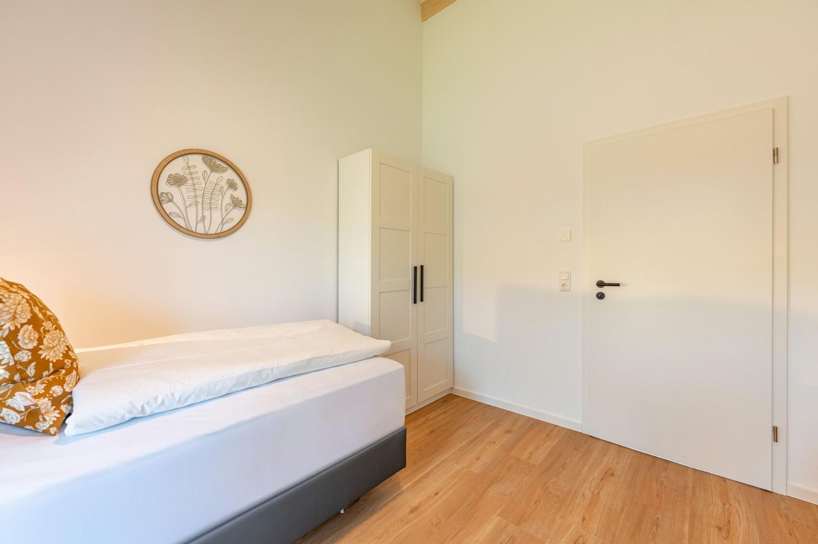 Schlafzimmer mit Bett, Kleiderschrank und Tür. Wanddekoration mit Pflanzenmotiv.
