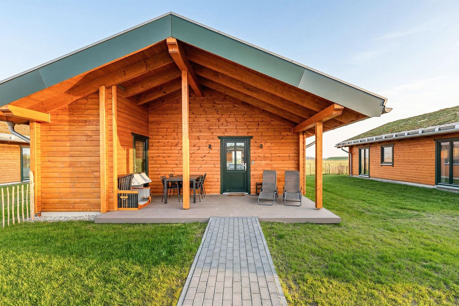 Holzhaus mit Terrasse, Tisch, Stühlen und Liegestühlen. Grasfläche und gepflasterter Weg.