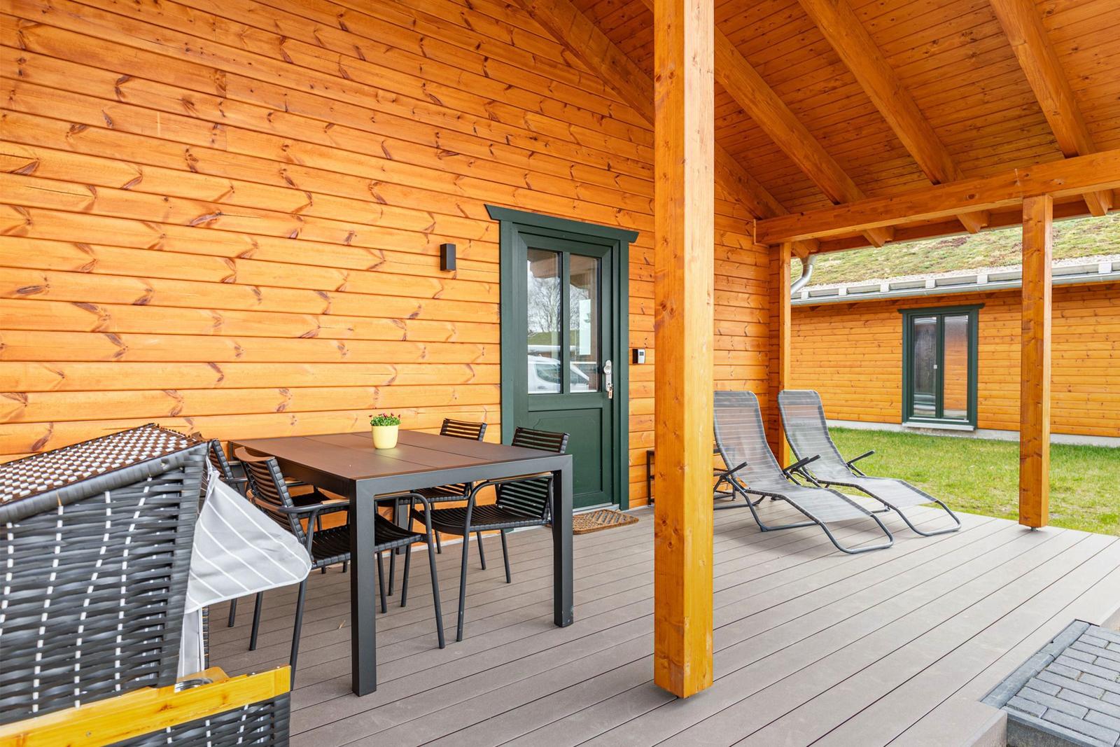 Terrasse mit Tisch, Stühlen und Liegen vor einem Holzhaus mit grüner Tür.