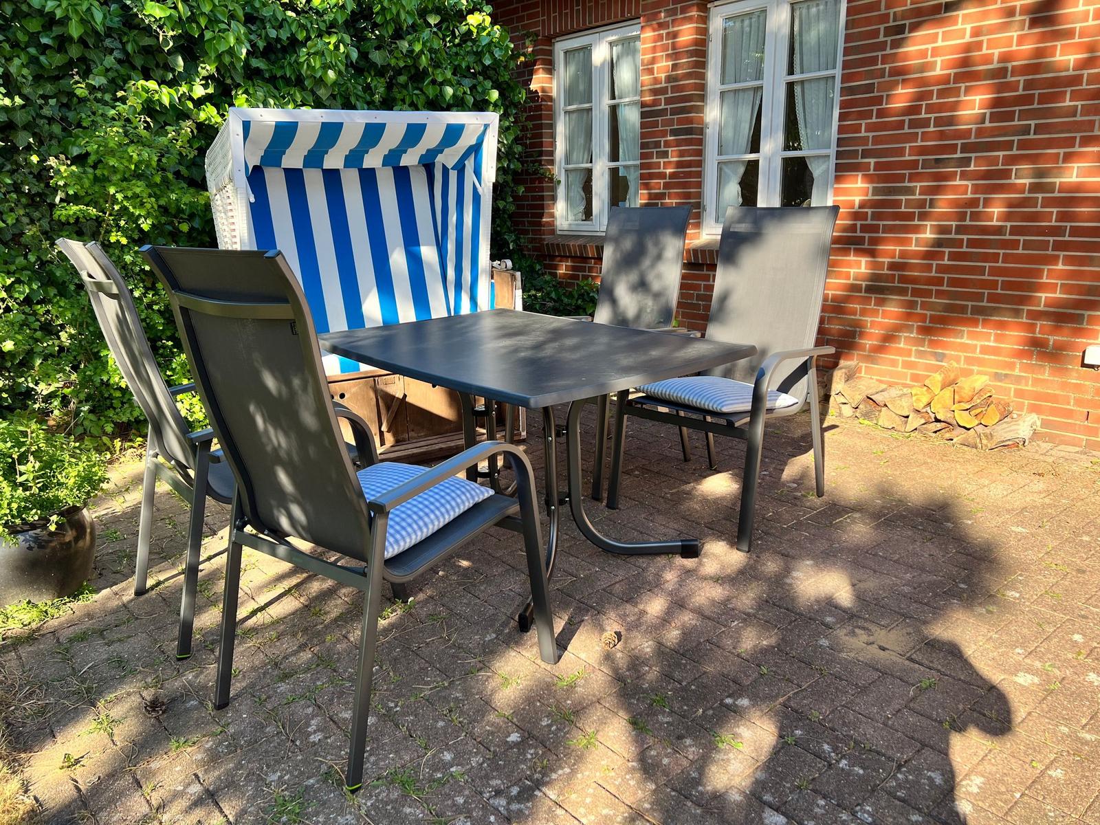 Terrasse mit Tisch, vier Stühlen und Strandkorb. Hintergrund: Backsteinhaus mit Fenstern.