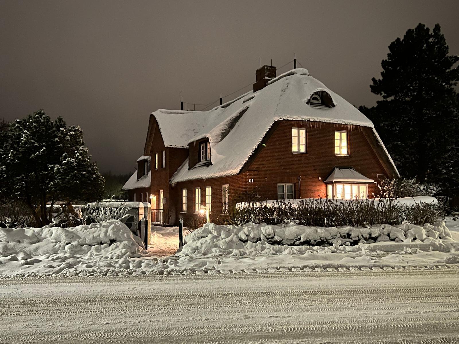 Schnees bedecktes Haus mit beleuchteten Fenstern und Schneehaufen vor dem Eingang.