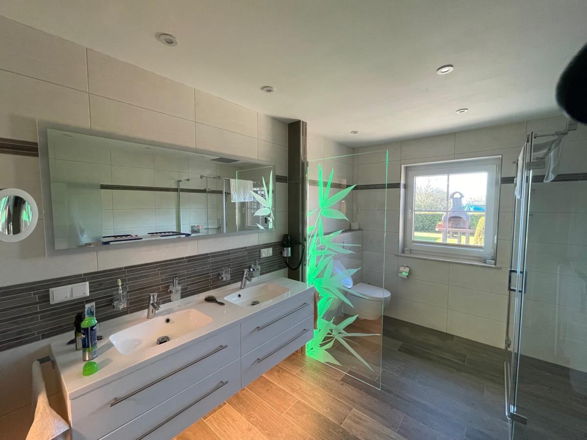 Bad mit Doppelwaschbecken, Glaswand und Toilette. Fenster zeigt Garten.