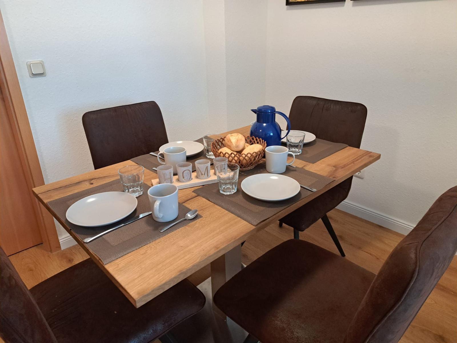 Holztisch mit vier Stühlen, Teller, Tassen und Brot im Korb.