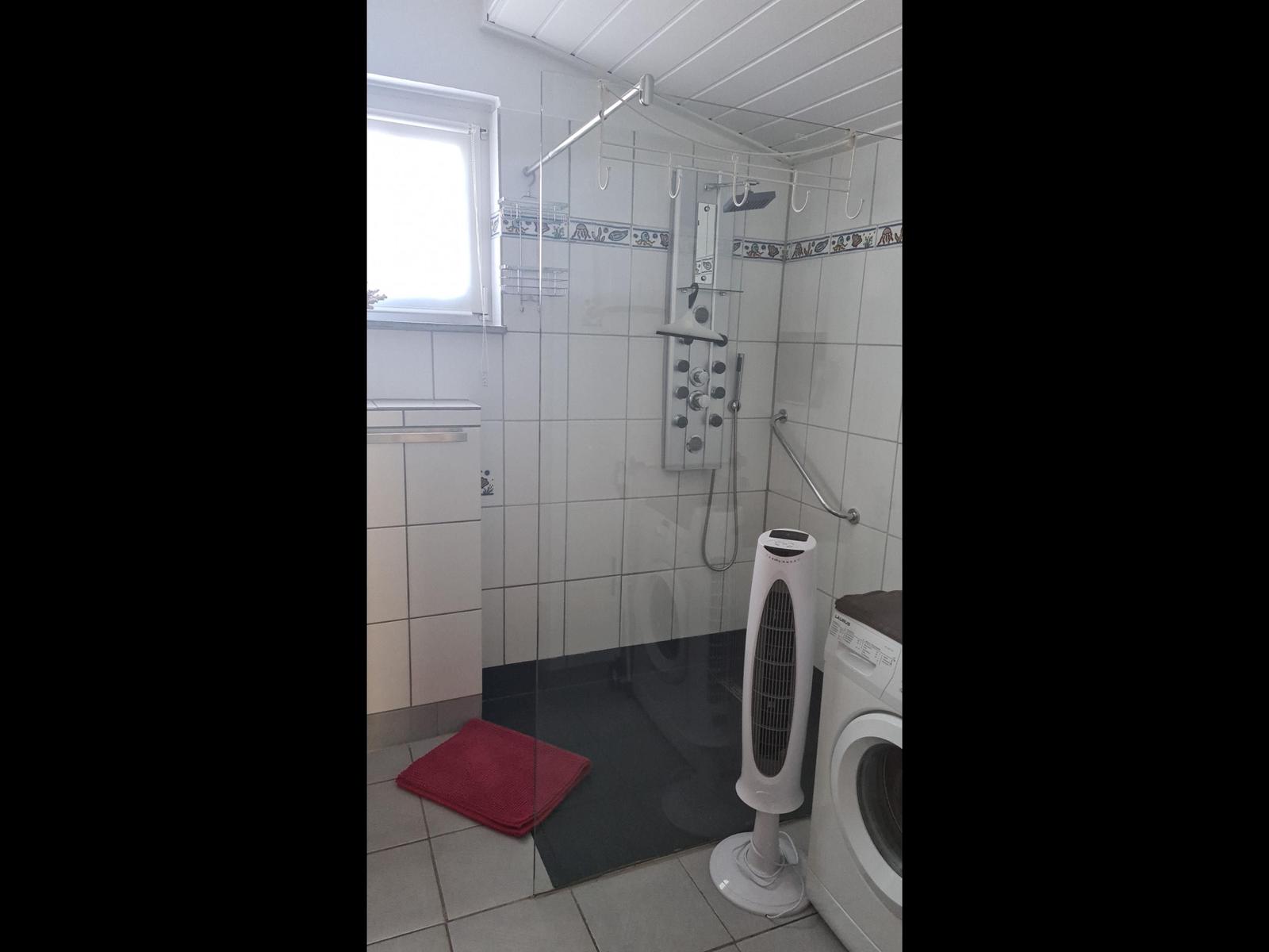 Badezimmer mit Dusche, Waschmaschine und Standventilator.