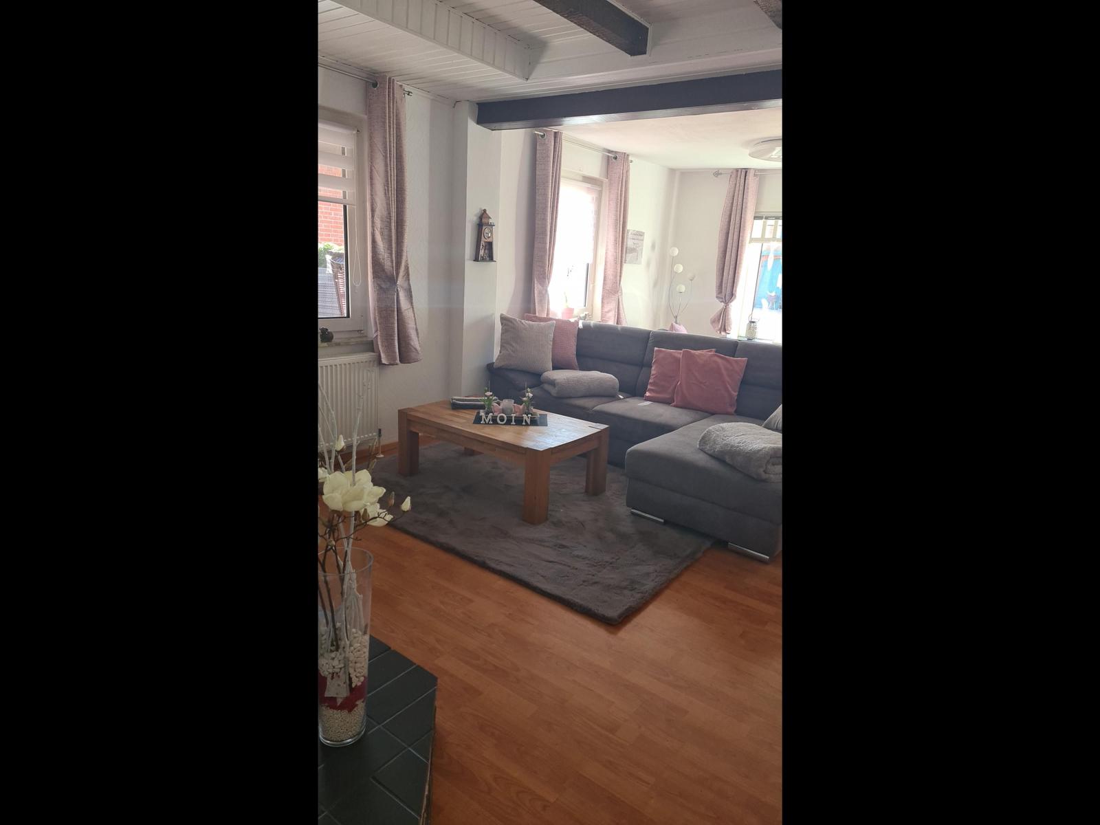 Wohnzimmer mit grauem Sofa, Holztisch und Holzboden. Fenster mit Vorhängen.