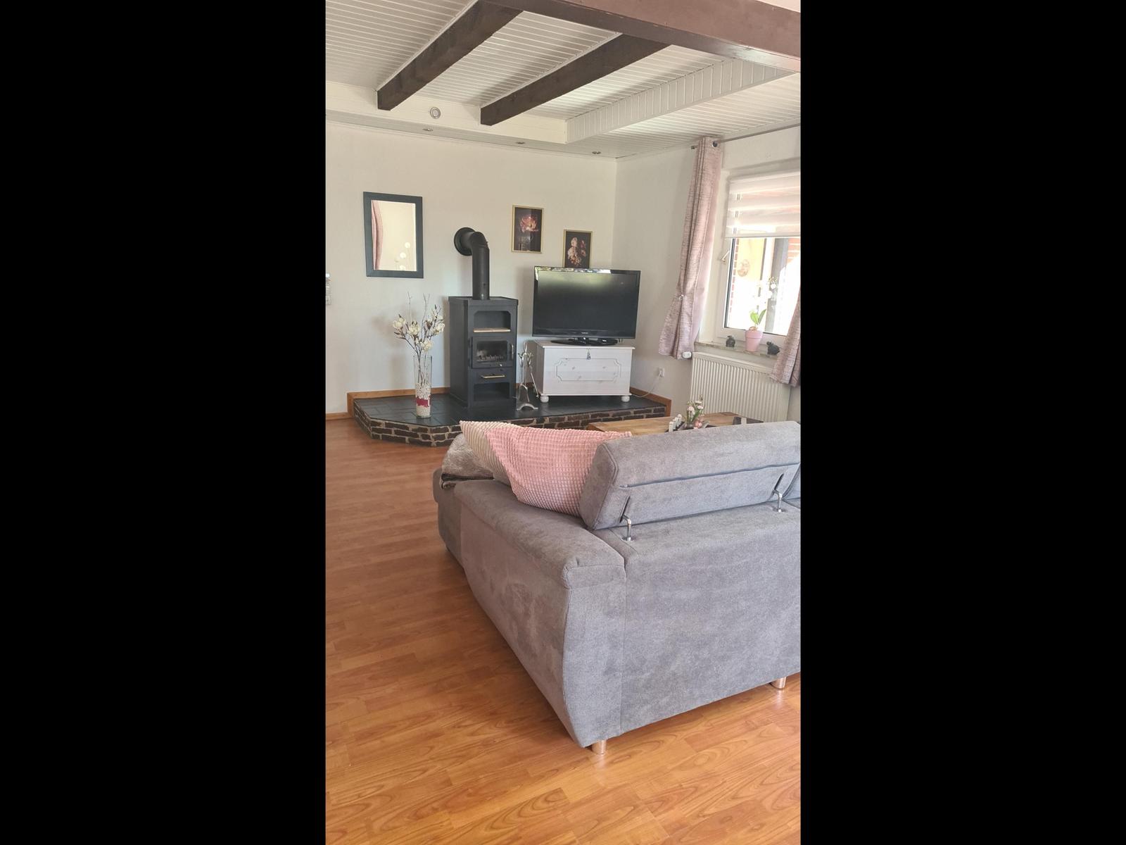 Wohnzimmer mit Sofa, Kaminofen und Fernseher. Holzfußboden und Balkendecke.