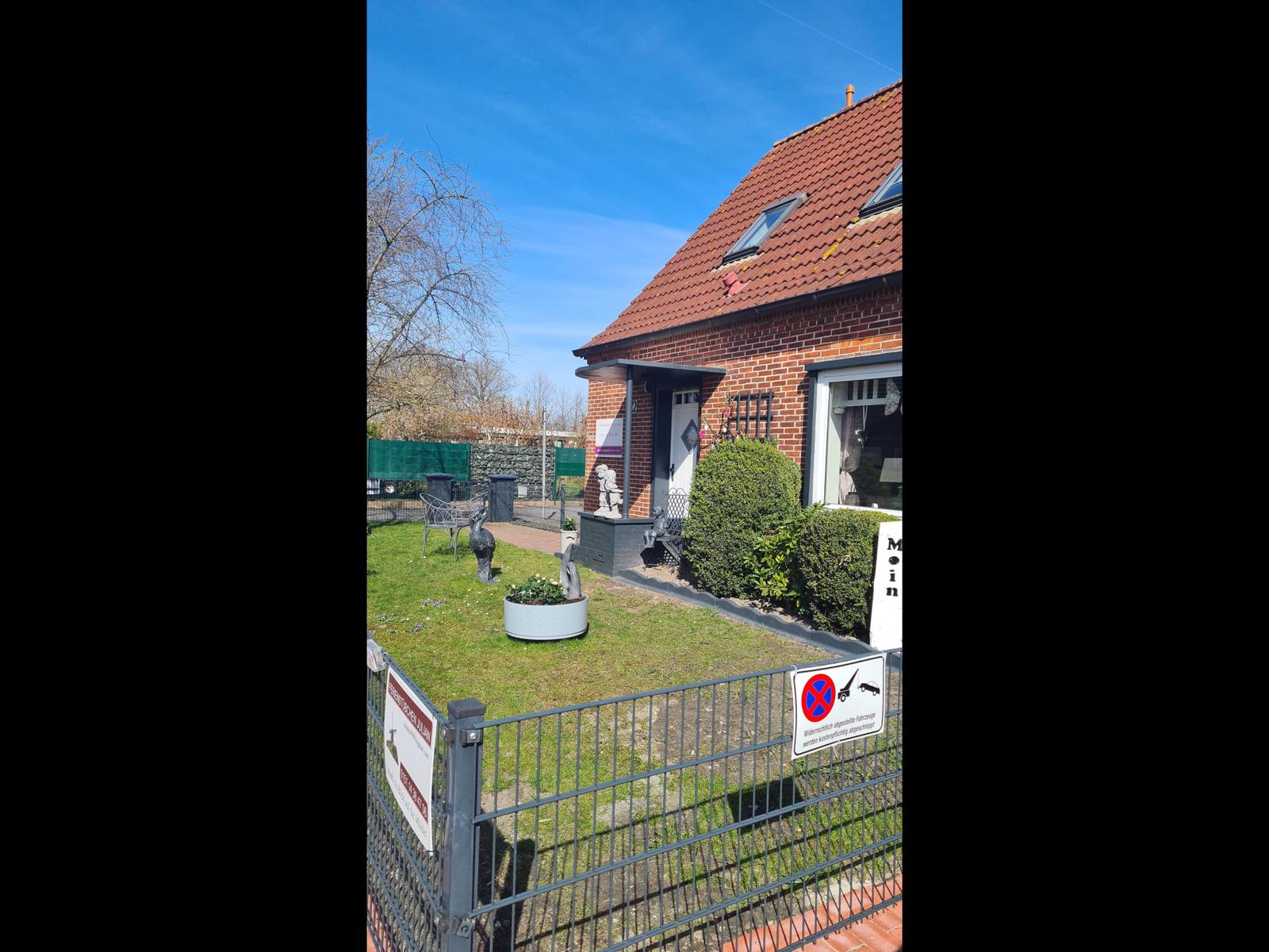 Haus mit rotem Dach, Garten mit Beete und Zaun. Vor dem Eingang steht eine Bank.