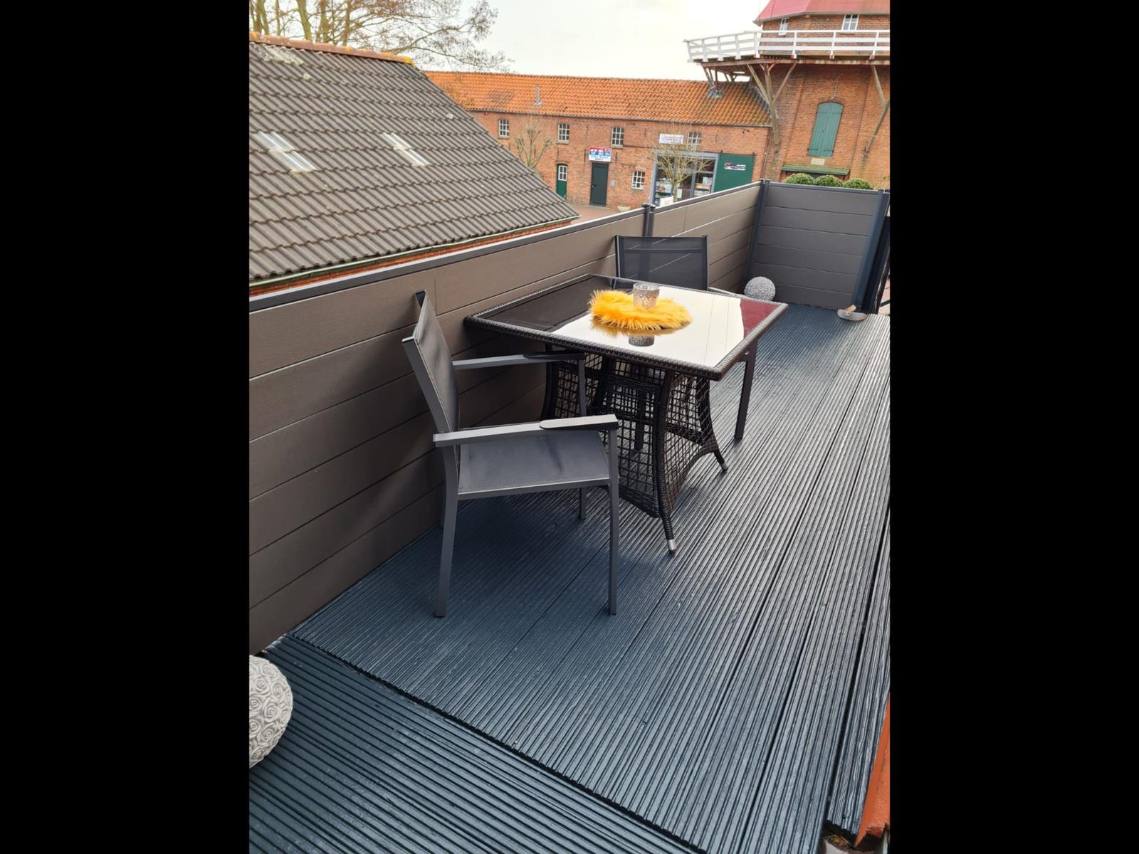 Terrasse mit Tisch und zwei Stühlen, dunkler Boden und Sicht auf Gebäude.