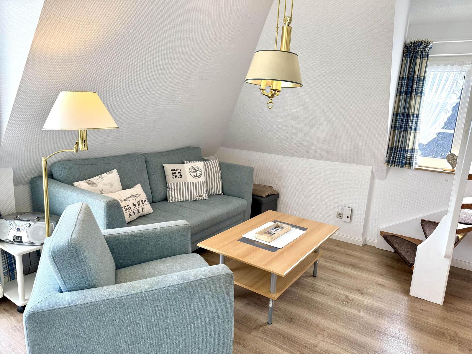 Living room with sofa, armchair, and table under a sloped ceiling. Window with curtains.