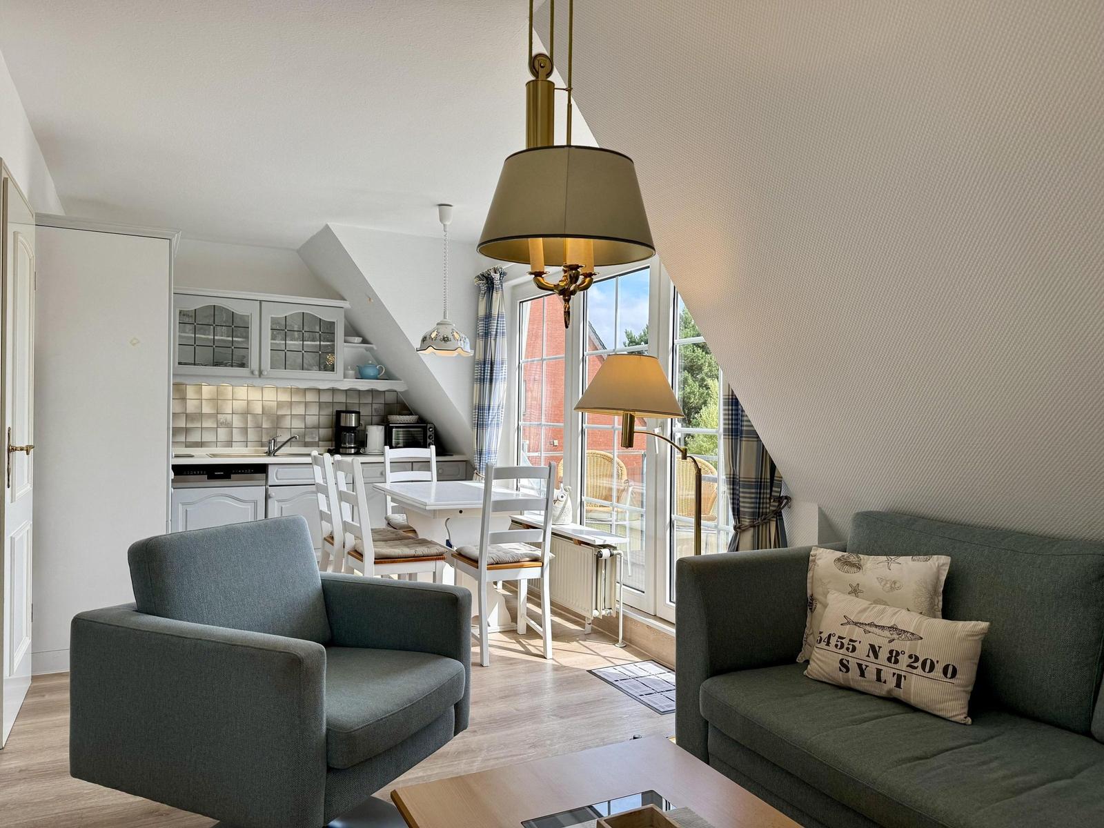 Living and dining area with kitchen under sloped ceiling. Sofa, armchair, and table are visible.