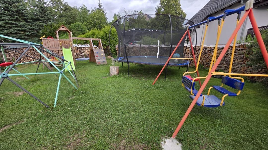 Hinterhof mit Spielplatz, Trampolin und Schaukel. Grasfläche und Holzstapel im Hintergrund.