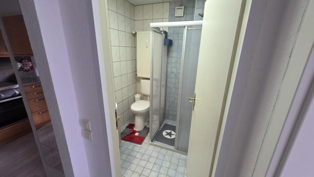 Badezimmer mit Dusche, Toilette und Fliesenboden. Küchenbereich ist teilweise sichtbar.