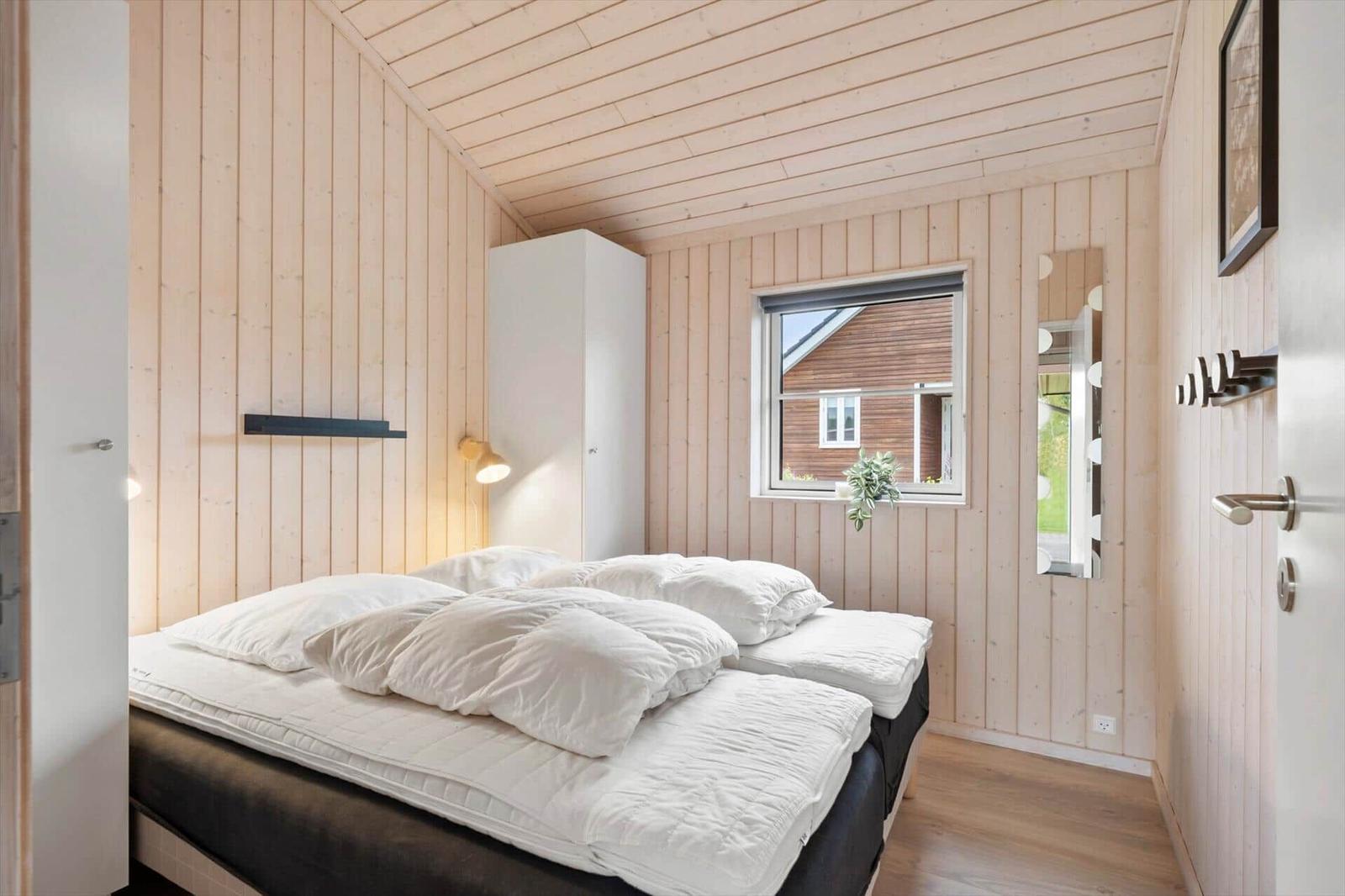 Bedroom with wooden wall cladding, bed with white pillows, and window view.