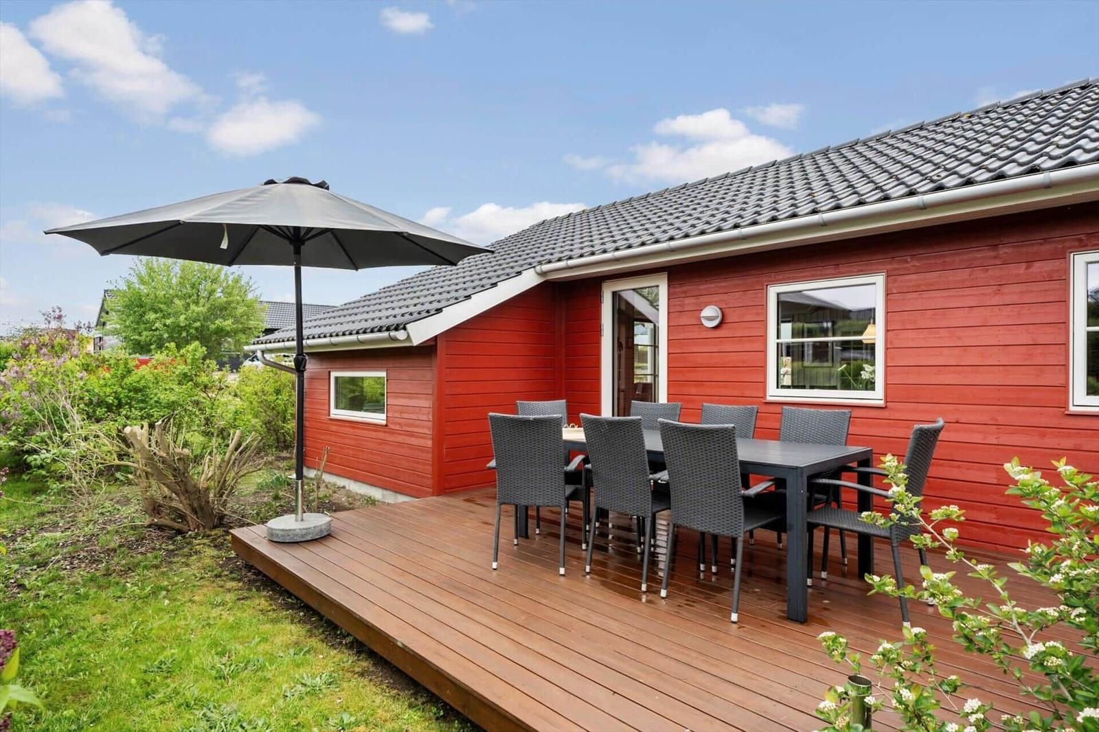 Red wooden house with terrace, table, chairs, and umbrella. Greenery and flowers surround the house.