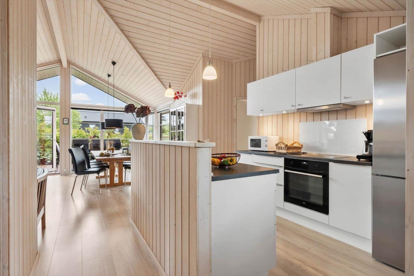 Kitchen with white cabinets, stainless steel fridge and oven. Wooden cladding and large window.