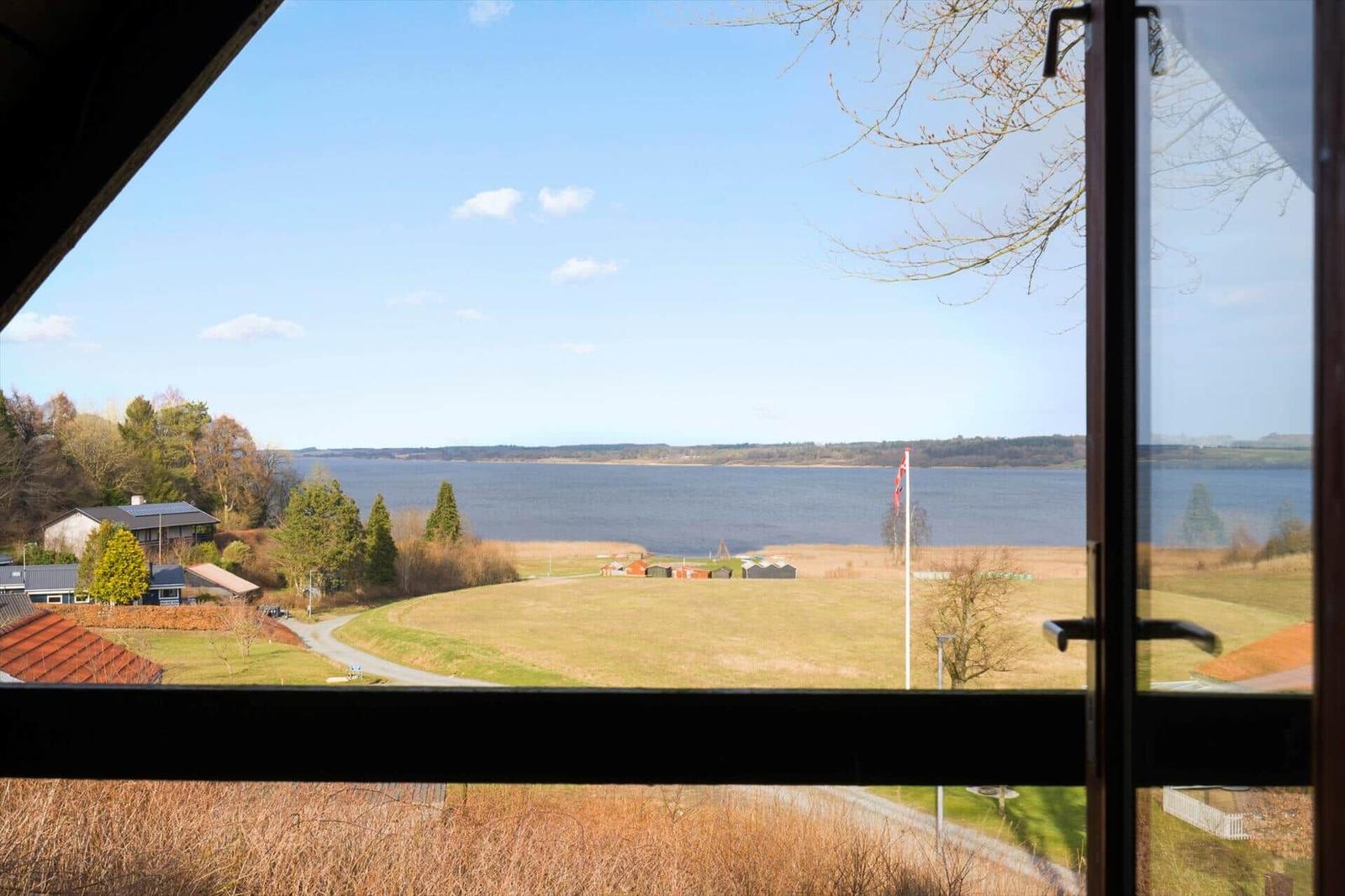 Blick aus dem Fenster auf einen See mit Grünflächen und einigen Häusern im Hintergrund.