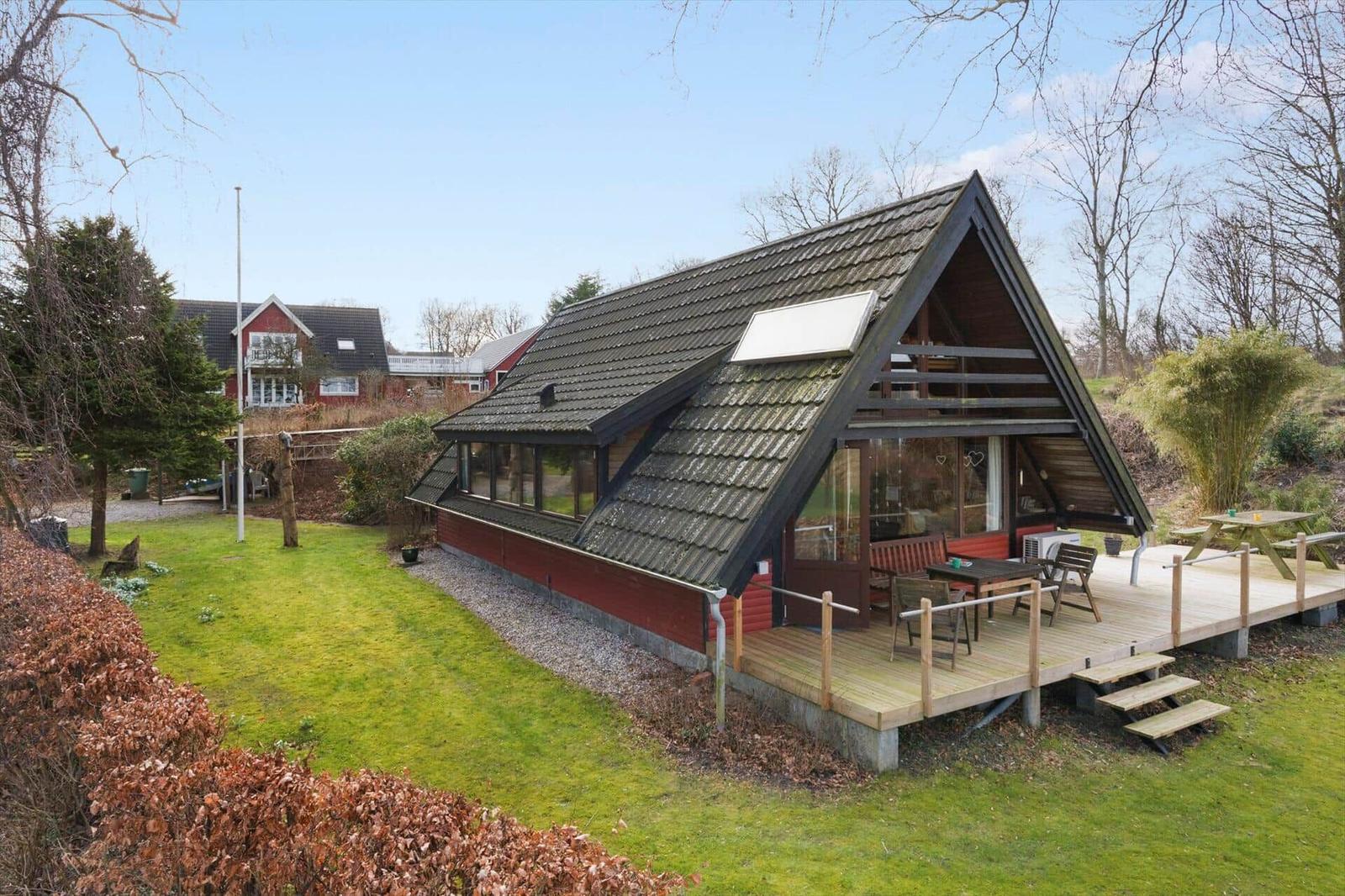 Holzhaus mit Terrasse, Garten und Blick auf Wald. Einrichtung im Inneren sichtbar.