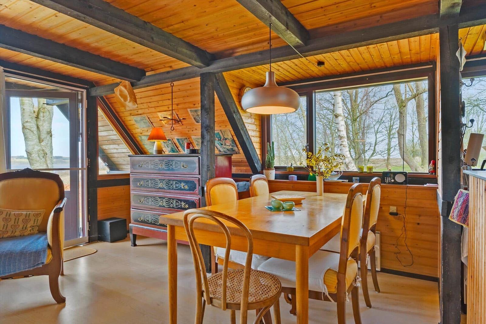 Esszimmer mit Holztisch, Stühlen und Blick auf Bäume. Holzwände und Deckenbalken.