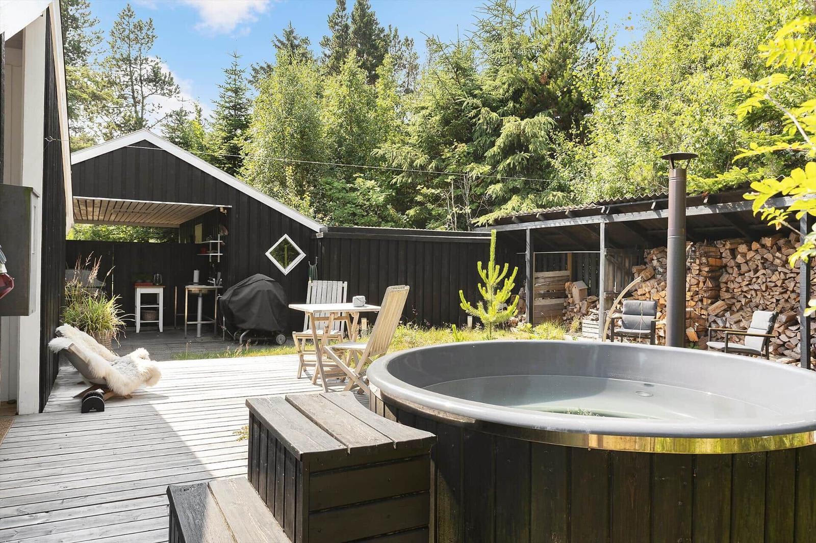 Backyard with hot tub, deck, and grill area. Surrounded by trees and woodpile.